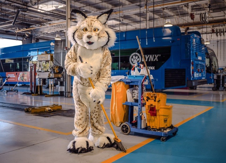 Person in a lynx mascot costume sweeping the floor inside of the lynx vehicle maintenance facility with cleaning supplies nearby.