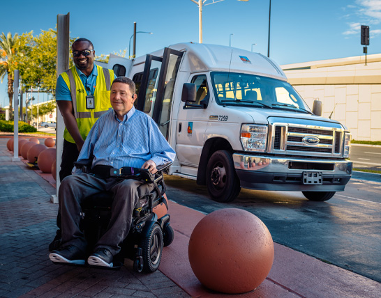 A man in a wheelchair is being assisted by a smiling transit worker near a white shuttle van on a sunny day.